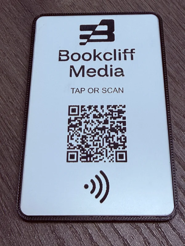A white acrylic Bookcliff Tap Card seated in a dark 3D-printed carrier on a wood surface, with a QR code and NFC tap symbol